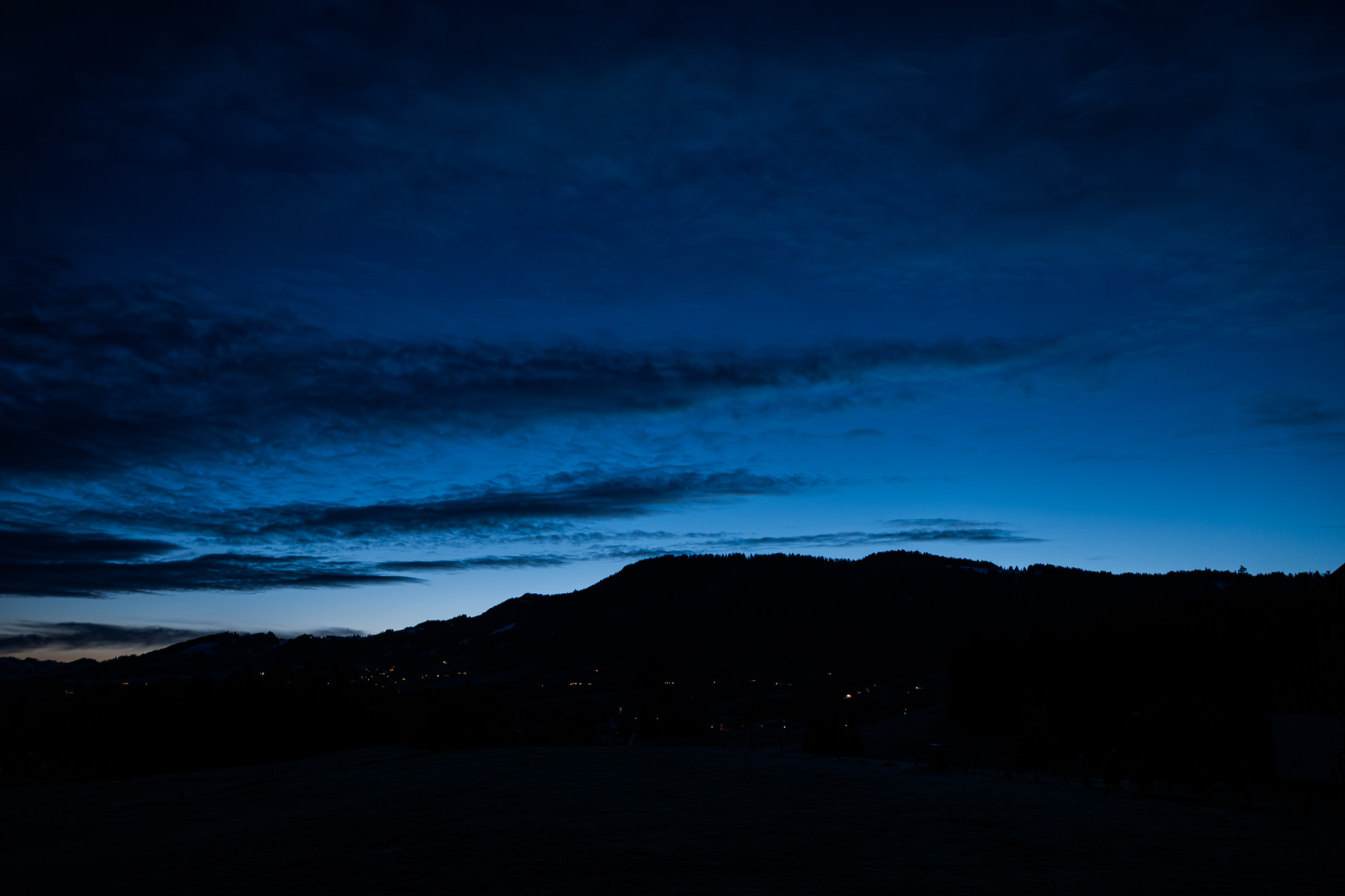 A serene twilight landscape featuring a dark mountain silhouette against a deep blue sky. Wispy clouds stretch across the horizon, illuminated by the fading light of dusk. In the foreground, scattered lights from a distant village dot the lower part of the image, adding a subtle warmth to the otherwise cool-toned scene.