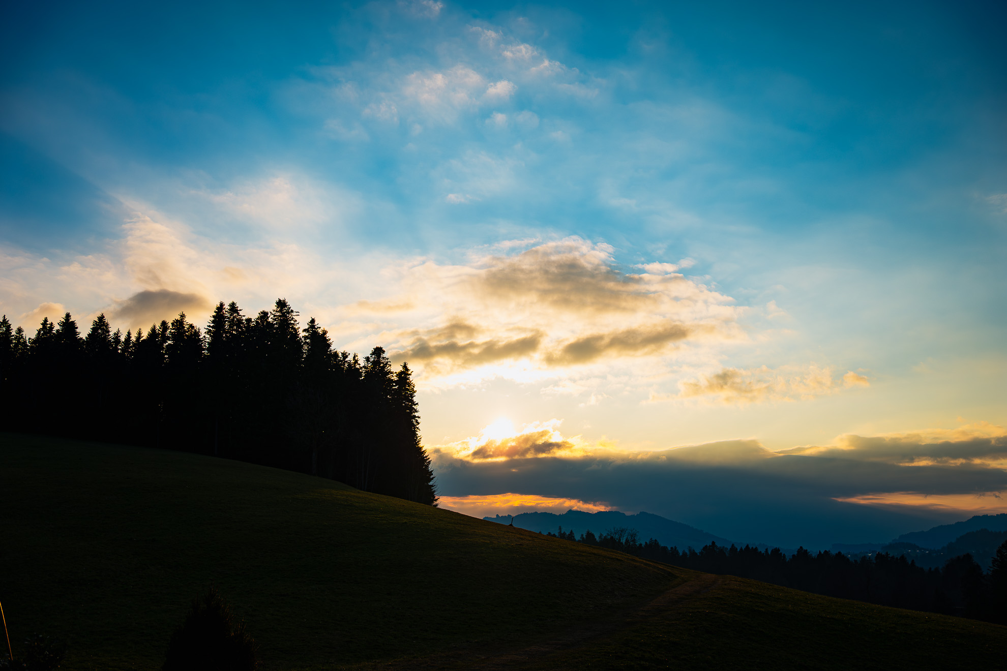 A scenic sunset over rolling hills, with the sun partially hidden behind clouds, casting a golden glow. The sky transitions from deep blue at the top to warm orange near the horizon. Silhouetted evergreen trees stand on a gentle hillside in the foreground, creating contrast against the bright sky. In the background, distant mountain ridges fade into a hazy blue, adding depth to the landscape.
